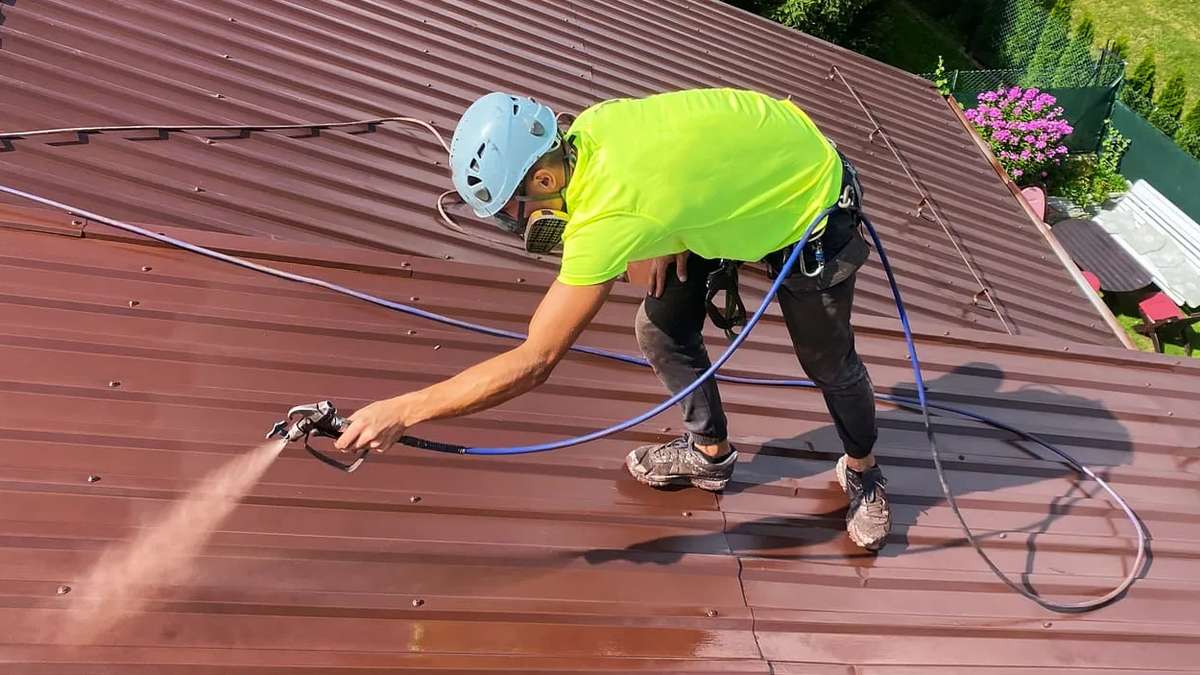 Un professionnel en harnais de sécurité pulvérisant une nouvelle couche de peinture sur un toit métallique décoloré.